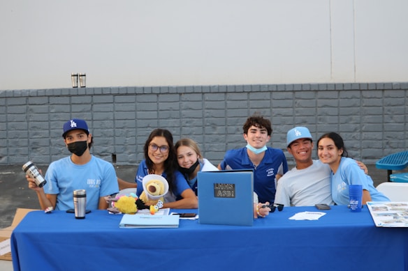 A group of six young individuals are sitting at a table covered with a blue tablecloth. They are smiling and appear to be part of a club or school event. Some are wearing blue shirts and hats with various logos. On the table, there are a few items such as a laptop, a stuffed toy, drink containers, and some papers.