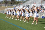 A group of cheerleaders in coordinated outfits perform a routine on a sports field. They are arranged in a line, each holding pom-poms raised above their heads. The bleachers in the background are partially filled with spectators. The scene takes place outdoors under clear skies.