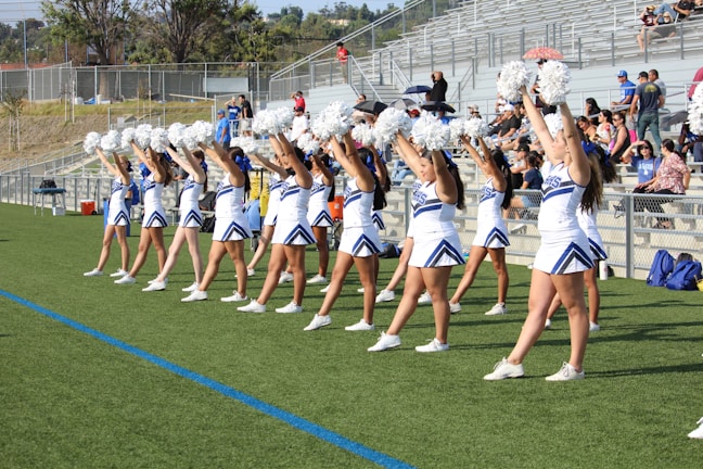 A group of cheerleaders in coordinated outfits perform a routine on a sports field. They are arranged in a line, each holding pom-poms raised above their heads. The bleachers in the background are partially filled with spectators. The scene takes place outdoors under clear skies.
