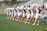 A group of cheerleaders in coordinated outfits perform a routine on a sports field. They are arranged in a line, each holding pom-poms raised above their heads. The bleachers in the background are partially filled with spectators. The scene takes place outdoors under clear skies.