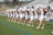 A group of cheerleaders in coordinated outfits perform a routine on a sports field. They are arranged in a line, each holding pom-poms raised above their heads. The bleachers in the background are partially filled with spectators. The scene takes place outdoors under clear skies.