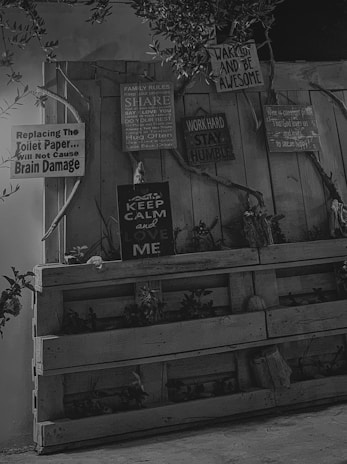 A monochrome image featuring several signs with humorous and motivational quotes attached to a wooden fence. The signs include messages like 'Replacing the toilet paper will not cause brain damage,' 'Keep calm and love me,' and 'Work hard, stay humble.' Some plants can be seen growing in wooden pallets at the bottom, and branches with leaves are hung around the setup.