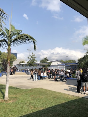 A group of people, primarily students, are gathered outside in a school setting. They are standing and sitting around blue picnic tables under the open sky and some scattered palm trees. The environment appears relaxed, with students socializing and enjoying the outdoor area. In the background, there are school buildings with white siding and blue accents, along with some trees and grass.