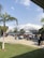 A group of people, primarily students, are gathered outside in a school setting. They are standing and sitting around blue picnic tables under the open sky and some scattered palm trees. The environment appears relaxed, with students socializing and enjoying the outdoor area. In the background, there are school buildings with white siding and blue accents, along with some trees and grass.