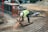 A construction worker wearing a high-visibility vest, hard hat, and protective gloves is bending over to use a tool that emits sparks on a metal grid. There are construction materials such as bricks, gravel, and bags of cement around the site, which is enclosed with metal barriers and red support frames. A wheelbarrow is also visible in the background.