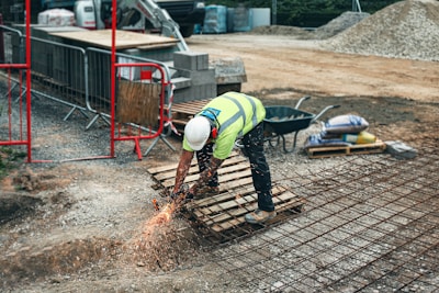 A construction worker wearing a high-visibility vest, hard hat, and protective gloves is bending over to use a tool that emits sparks on a metal grid. There are construction materials such as bricks, gravel, and bags of cement around the site, which is enclosed with metal barriers and red support frames. A wheelbarrow is also visible in the background.