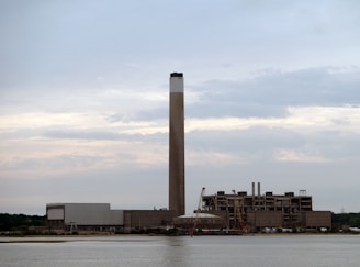 A large industrial facility featuring a tall smokestack stands near a body of water. The structure is expansive with several sections and appears to be made of concrete. Cranes are positioned near the building, suggesting ongoing construction or maintenance work. The sky is overcast with gray and light blue clouds.