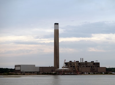 A large industrial facility featuring a tall smokestack stands near a body of water. The structure is expansive with several sections and appears to be made of concrete. Cranes are positioned near the building, suggesting ongoing construction or maintenance work. The sky is overcast with gray and light blue clouds.