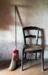 A wooden chair with a woven seat is positioned against a textured wall. Next to it is a broom with a red handle and straw bristles. The floor is made of terracotta tiles, and soft warm light creates subtle shadows.