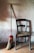 A wooden chair with a woven seat is positioned against a textured wall. Next to it is a broom with a red handle and straw bristles. The floor is made of terracotta tiles, and soft warm light creates subtle shadows.