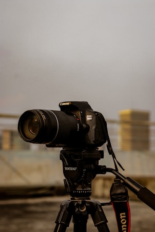 A Canon DSLR camera mounted on a Yunteng tripod is prominently displayed against a blurred urban background. The camera is facing forward and the image captures its lens and body in detail, emphasizing the professional equipment.