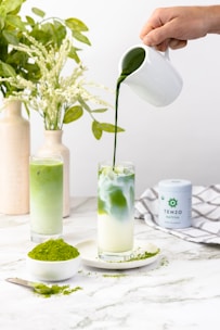A hand pours green matcha tea from a white pitcher into a glass filled with milk, creating a marbled effect. Beside it, there is another glass with a green matcha drink. A spoon and small plate with matcha powder are on the table, surrounded by a clean, white marble surface. A container of matcha powder and decorative vases with green leaves are in the background.
