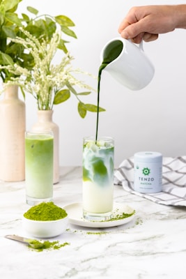 A hand pours green matcha tea from a white pitcher into a glass filled with milk, creating a marbled effect. Beside it, there is another glass with a green matcha drink. A spoon and small plate with matcha powder are on the table, surrounded by a clean, white marble surface. A container of matcha powder and decorative vases with green leaves are in the background.