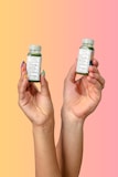 Two hands are holding small green beverage bottles labeled 'magic mind' against a gradient background that transitions from yellow to pink. The bottles have labels indicating ingredients such as matcha and honey. The nails on the hands are painted in different pastel colors.
