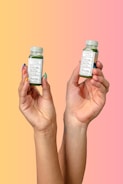Two hands are holding small green beverage bottles labeled 'magic mind' against a gradient background that transitions from yellow to pink. The bottles have labels indicating ingredients such as matcha and honey. The nails on the hands are painted in different pastel colors.