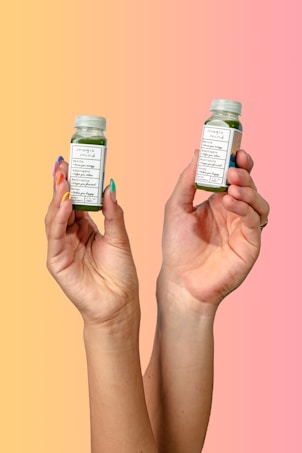 Two hands are holding small green beverage bottles labeled 'magic mind' against a gradient background that transitions from yellow to pink. The bottles have labels indicating ingredients such as matcha and honey. The nails on the hands are painted in different pastel colors.