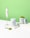 A clean and minimalist composition featuring containers of Tenzo Matcha green tea powder alongside an electric whisk. The image showcases one container opened with matcha powder spilling out on a white surface and a scooping spoon nearby. The background is a vibrant green, complementing the green tea theme.