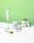 A clean and minimalist composition featuring containers of Tenzo Matcha green tea powder alongside an electric whisk. The image showcases one container opened with matcha powder spilling out on a white surface and a scooping spoon nearby. The background is a vibrant green, complementing the green tea theme.