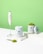 A clean and minimalist composition featuring containers of Tenzo Matcha green tea powder alongside an electric whisk. The image showcases one container opened with matcha powder spilling out on a white surface and a scooping spoon nearby. The background is a vibrant green, complementing the green tea theme.