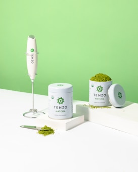 A clean and minimalist composition featuring containers of Tenzo Matcha green tea powder alongside an electric whisk. The image showcases one container opened with matcha powder spilling out on a white surface and a scooping spoon nearby. The background is a vibrant green, complementing the green tea theme.
