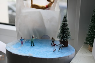 A miniature scene with small figurines ice skating on a circular patch of blue ice. Surrounded by snow and a small, snow-capped pine tree, the figurines are dressed in colorful winter clothing with one pair appearing to skate together. The setting is indoors, near a window, as part of a decorative display.