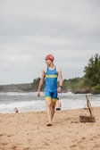 A person wearing a blue and yellow triathlon suit and a red swim cap walks barefoot on a sandy beach. They are wearing sunglasses and a wristwatch. The ocean waves are visible in the background, along with a few other people relaxing on the beach. Trees and hilly terrain are seen in the distance under a cloudy sky.