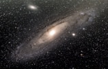 The image depicts a large spiral galaxy with a bright central nucleus surrounded by a diffuse halo of stars and dust. Numerous stars are scattered across the scene, twinkling against the dark backdrop of space. There are distinct spiral arms wrapping around the galaxy's core, creating a sense of depth and structure.