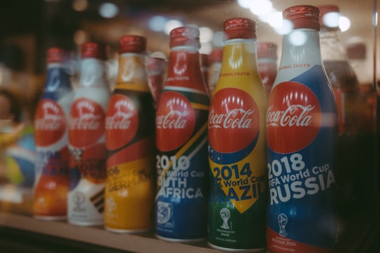 A row of Coca-Cola bottles with various designs commemorating the FIFA World Cup for different years, including 1998 in France, 2002 in South Korea and Japan, 2006 in Germany, 2010 in South Africa, 2014 in Brazil, and 2018 in Russia. Each bottle features vibrant colors and branding associated with the respective tournament.