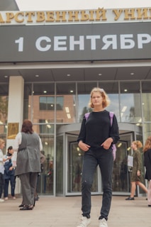 A person stands confidently in front of a building entrance with a sign that reads '1 September' in Russian. Several people are gathered and interacting in the background. The person in the foreground is wearing a black outfit and carrying a purple backpack.