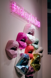 A neon pink sign with the hashtag #makeupkitchen hangs above a series of multicolored, geometric 3D sculptures of lips mounted on a wall. The lips are crafted in various shades, including pink, red, blue, and green, displaying an array of vibrant and metallic textures.