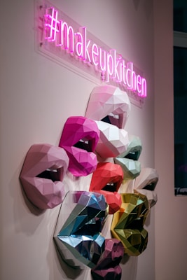 A neon pink sign with the hashtag #makeupkitchen hangs above a series of multicolored, geometric 3D sculptures of lips mounted on a wall. The lips are crafted in various shades, including pink, red, blue, and green, displaying an array of vibrant and metallic textures.