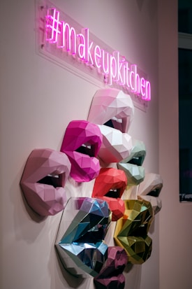 A neon pink sign with the hashtag #makeupkitchen hangs above a series of multicolored, geometric 3D sculptures of lips mounted on a wall. The lips are crafted in various shades, including pink, red, blue, and green, displaying an array of vibrant and metallic textures.