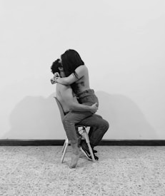 A couple is engaged in an intimate moment, sitting on a single chair against a plain wall in a minimalist room. The woman is straddling the man, and both appear to be shirtless, with the woman's back showing a tattoo. The floor is tiled and the lighting creates shadows on the wall behind them.