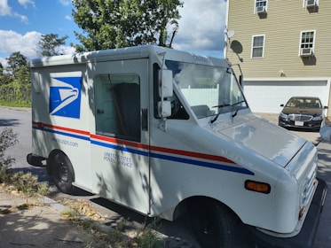 A United States Postal Service (USPS) delivery truck is parked on a residential street. The truck is white with the USPS logo and red and blue stripes. In the background, there is a light-colored house with a garage and a dark car parked in the driveway. Trees and a clear blue sky with clouds are visible.