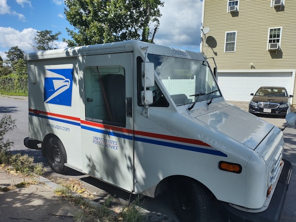 A United States Postal Service (USPS) delivery truck is parked on a residential street. The truck is white with the USPS logo and red and blue stripes. In the background, there is a light-colored house with a garage and a dark car parked in the driveway. Trees and a clear blue sky with clouds are visible.