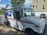 A United States Postal Service (USPS) delivery truck is parked on a residential street. The truck is white with the USPS logo and red and blue stripes. In the background, there is a light-colored house with a garage and a dark car parked in the driveway. Trees and a clear blue sky with clouds are visible.