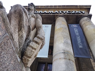 A large stone statue of a man is prominently displayed in front of a building with tall columns and banners hanging from them. The building features architectural elements typical of grand structures, with the word 'Dramatyczny' visible on the frieze. The statue appears to be intricately carved, showing detailed textures.