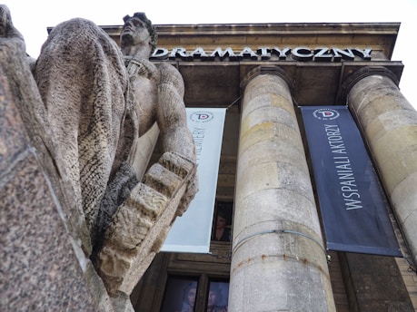 A large stone statue of a man is prominently displayed in front of a building with tall columns and banners hanging from them. The building features architectural elements typical of grand structures, with the word 'Dramatyczny' visible on the frieze. The statue appears to be intricately carved, showing detailed textures.