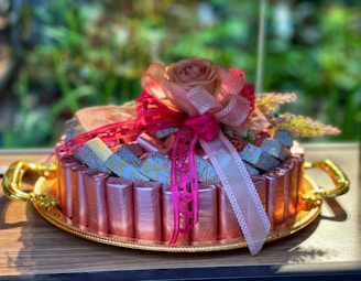 An elegantly decorated arrangement of wrapped candies or chocolates on a golden tray. The treats are surrounded by intricate ribbons and topped with an artificial peach-colored rose. The setting is on a wooden surface with a blurred green background, suggesting an indoor location with natural plants.