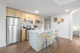 A modern kitchen with a minimalist design featuring stainless steel appliances, including a refrigerator, oven, and microwave. Light wood cabinets complement the sleek white countertops. A central island with a sink has two wooden barstools with metal legs. Decorative plates hang on a white wall, and wood flooring adds warmth to the space.