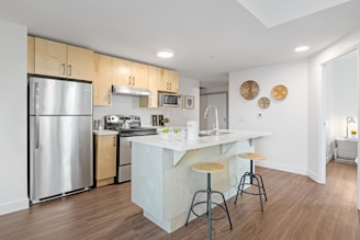 A modern kitchen with a minimalist design featuring stainless steel appliances, including a refrigerator, oven, and microwave. Light wood cabinets complement the sleek white countertops. A central island with a sink has two wooden barstools with metal legs. Decorative plates hang on a white wall, and wood flooring adds warmth to the space.