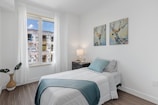 A minimalistic bedroom with a single bed covered in white linens and a blue throw. Two paintings depicting deer hang on the wall above the bed. A nightstand next to the bed holds a lamp and some decorative items. A large window with sheer curtains allows light to enter, with the view of a building under construction outside. A plant in a wicker pot is placed on the floor beside the window.
