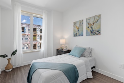 A minimalistic bedroom with a single bed covered in white linens and a blue throw. Two paintings depicting deer hang on the wall above the bed. A nightstand next to the bed holds a lamp and some decorative items. A large window with sheer curtains allows light to enter, with the view of a building under construction outside. A plant in a wicker pot is placed on the floor beside the window.