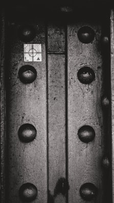 A close-up view of a metallic surface with large rivets or bolts. There is a small grid-marked paper or sticker with a target-like symbol and the letters L and A on it. The texture of the metal is rough and worn.