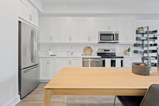A modern kitchen features white cabinetry, a stainless steel refrigerator, oven, and microwave. A wooden dining table is in the foreground with a decorative bowl on it, and open shelves display glassware and a small plant.