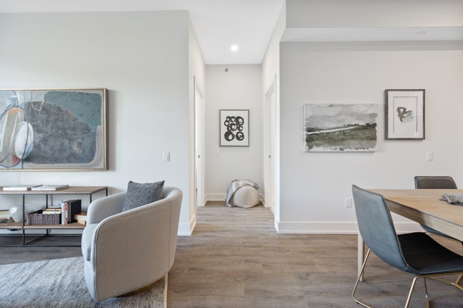 A modern living space with minimalistic and contemporary design elements. The room features a large abstract painting on the wall above a console table with books and decorative items. A soft grey armchair with a patterned pillow is placed near a light wood dining table and black chairs. The flooring is wooden with a grey-toned rug partially visible. Additional artwork is displayed on an adjacent wall, and a woven basket can be seen down a hallway.