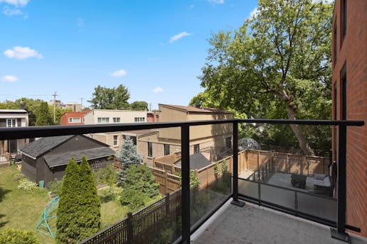 A view from a balcony with a transparent glass railing overlooking a backyard. The scene includes several residential buildings, a grass lawn with small trees and shrubs, a black shed, and a patio area with outdoor furniture. The sky is clear with a few clouds and large trees surround the area, providing greenery and shade.