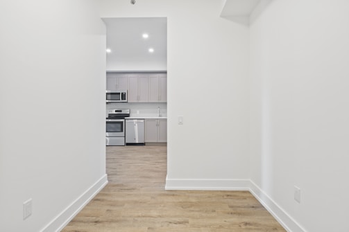 A minimalistic kitchen viewed through a narrow hallway, featuring modern gray cabinetry, stainless steel appliances including a stove and microwave, with a wooden floor and bright overhead lighting.