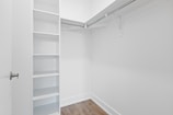 An empty walk-in closet with white walls, featuring built-in shelves on the left and a hanging rod extending along two walls. The floor is finished with a light brown wood texture, and there is a closed door with a metallic handle at the entrance.