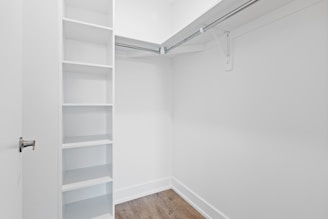 An empty walk-in closet with white walls, featuring built-in shelves on the left and a hanging rod extending along two walls. The floor is finished with a light brown wood texture, and there is a closed door with a metallic handle at the entrance.